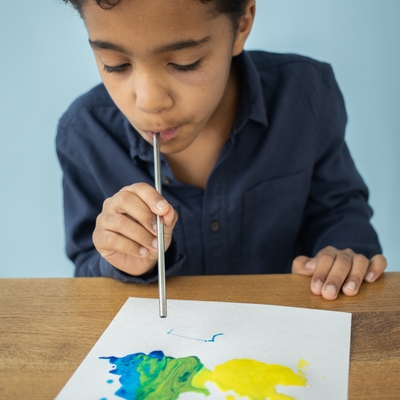 A boy blowing in a straw to move paint on a piece of paper.