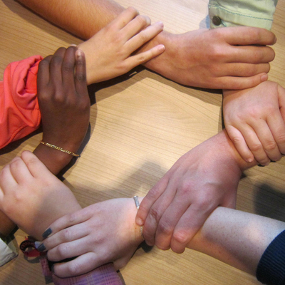 Six people from diverse races form a circle of trust using their hands, with one person's hand holding the wrist of another person. 
