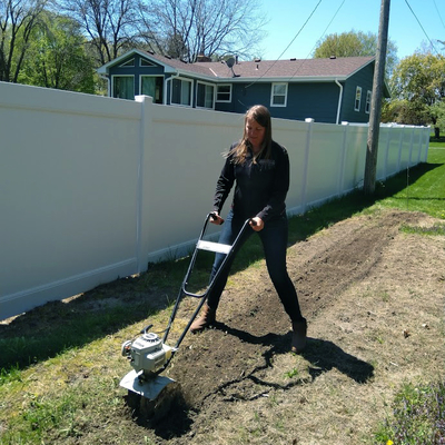 Woman using a rototiller to till long strip of ground in a yard.