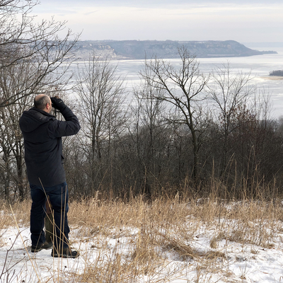 A man looks through binoculars at a river valley in winter.