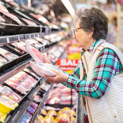 A senior woman in a grocery store compares prices on two packages of meat.