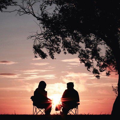 Silhouettes of two people at sunrise sitting on chairs outside near a tree.