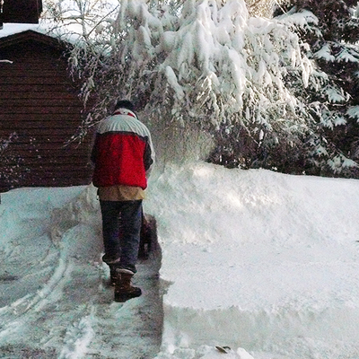 Man in winter clothing using a snowblower to remove snow from a driveway and blowing it onto a birch tree and two large spruce trees.