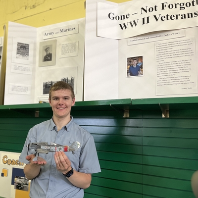 Zach W. holding his plane exhibit in front of a display board titled, "Gone - Not Forgotten - WWII Veterans."