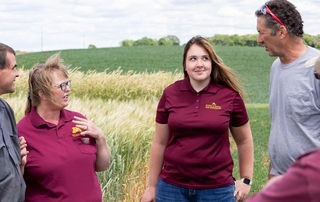 The three people in view are in a field of grain. Abby and Colleen are wearing Extension polo shirts. Abby is looking at the farmer, Ron while he talks.