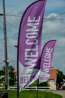 Two large purple flags that read "welcome a place to belong" are near a road leading into a town.