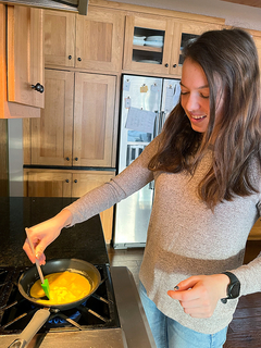 A teenage girl stands at a stove cooking scrambled eggs.