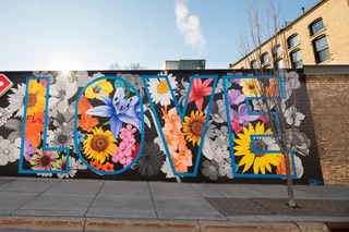 A community business that supports diversity and inclusion has a large mural with the word love bordered in blue and filled in with various colorful flowers. The letters are on top of a black and white floral background, which shows the contrast between the bright colors on top and the neutral ones below.