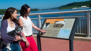 Two young women, one holding a small dog, reading an outdoor-historical landmark sign.